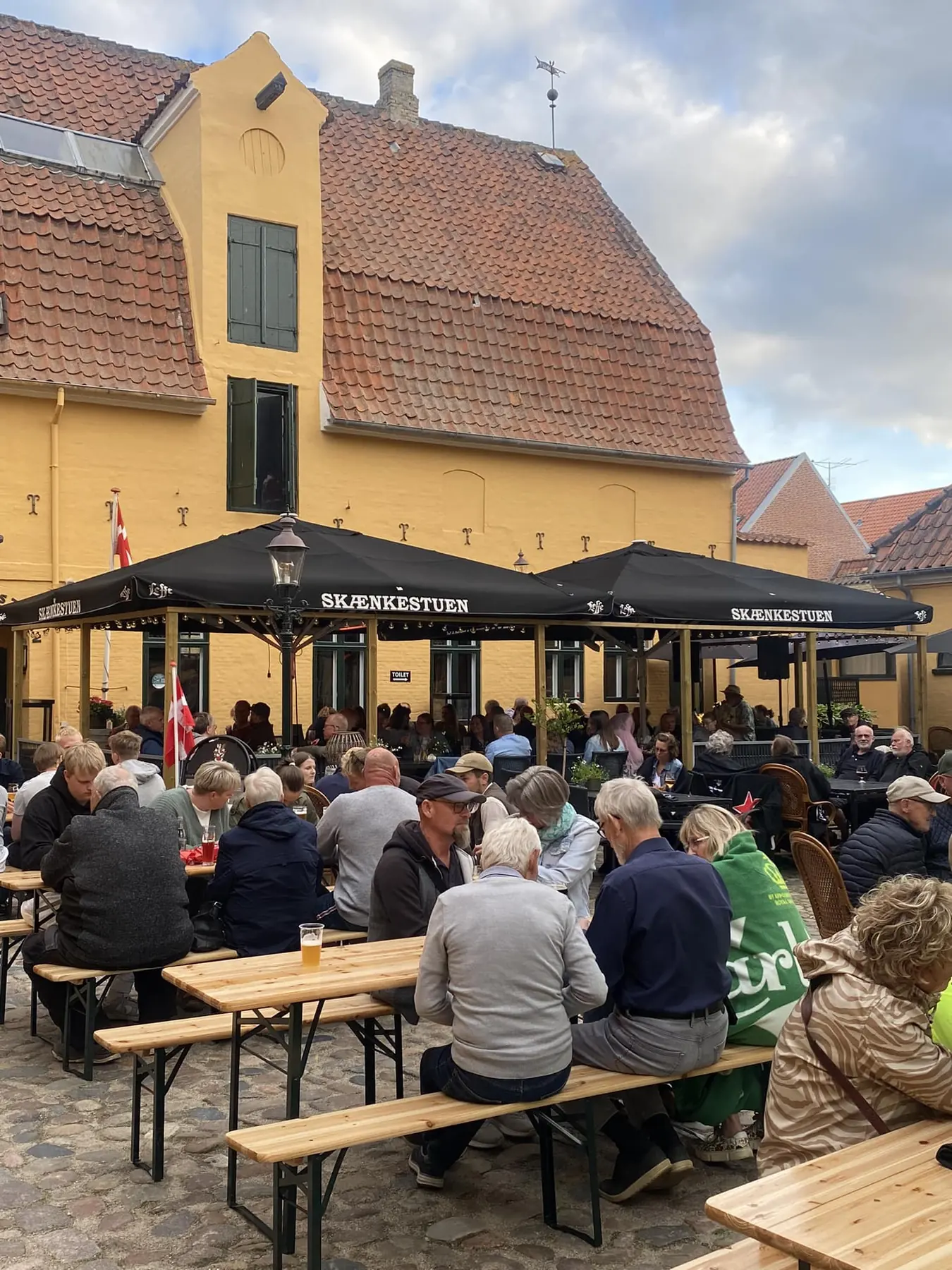 Fyldt gårdsplads med bænke foran de gule bygninger i Plums Gård